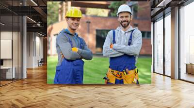 Experts in remodelling. Portrait of two handsome young male engineers in hard hats smiling at camera, posing outdoors with arms crossed while working on cottage construction Wall mural