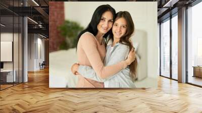 Forever best friend. Happy latin mother and daughter hugging each other, smiling at camera, looking affectionate while posing together indoors Wall mural