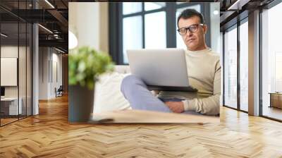 Freelance worker. Portrait of latin middle aged business man with eyeglasses working at home on some project, sitting on a sofa with his laptop Wall mural