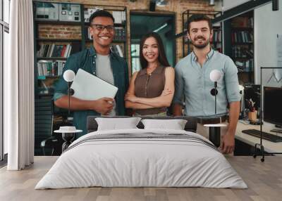 Happy to work together. Group of three cheerful young people in casual wear looking at camera with smile while standing in modern office Wall mural