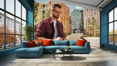 I can work from anywhere. Young handsome man with stubble in casual clothes and eyeglasses working on laptop while sitting on the bench near his bicycle Wall mural