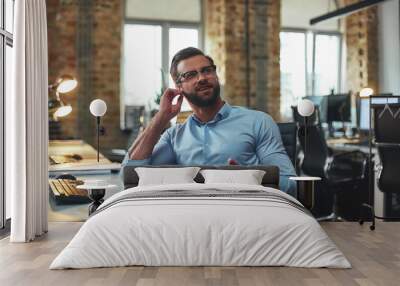 I don't hear you. Portrait of young bearded man in eyeglasses and headphones talking with client while sitting in the modern office Wall mural