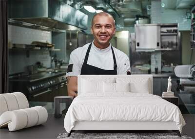 I love my work Cheerful young chef in apron keeping tattooed arms crossed and smiling while standing in a restaurant kitchen Wall mural