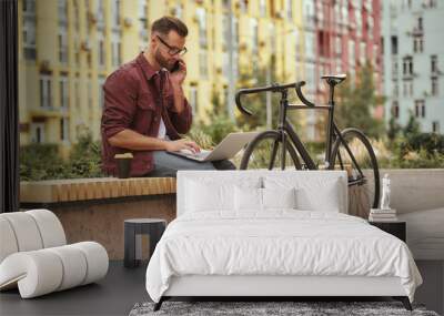 Important talk with client. Handsome and young man with stubble in casual clothes working on laptop and talking by phone while sitting on the bench near his bicycle Wall mural