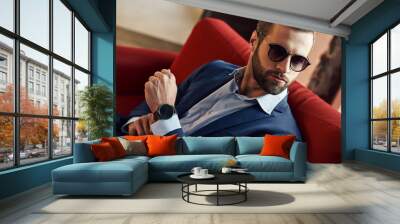 Looking just perfect...Young and sexy bearded businessman in sunglasses is looking away while sitting on office sofa Wall mural