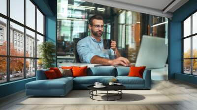 Morning coffee. Portrait of young and successful bearded man in eyeglasses holding cup of coffee and working with laptop while sitting at his working place Wall mural