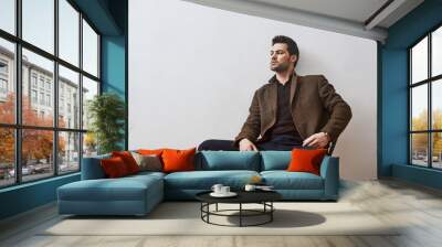 Need a rest. Pensive brunette man in brown jacket and blue trousers sitting on the chair and looking away over white background Wall mural