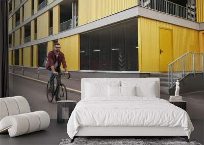 New bike. Handsome man with stubble in casual clothes riding on his bicycle along a road against yellow building Wall mural