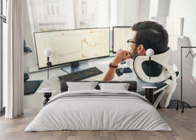 No time to relax. Back view of professional trader working with graph and financial reports on computer screens while sitting in his modern office. Wall mural