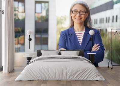 Portrait of a beautiful happy mature business woman in eyeglasses and classic wear keeping arms crossed and smiling at camera while standing against office building outdoors Wall mural