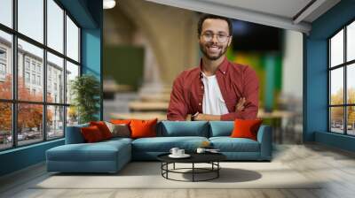 Portrait of a young cheerful caucasian male office worker looking at camera and smiling, keeping arms crossed while standing in the modern coworking space Wall mural