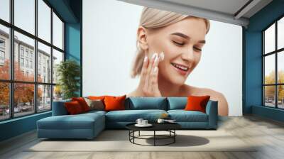 Portrait of cute blonde woman spreading cream on her face and smiling while standing against grey background. Skin care Wall mural