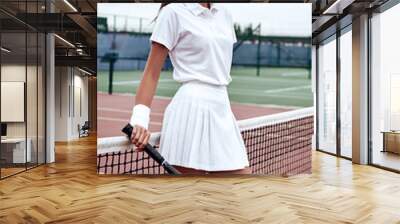 Portrait of fashionable woman in white clothing and cap with tennis racket posing at tennis net on court. Sports Fashion Wall mural