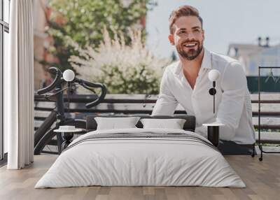 Ready to go. Young smiling man sitting on a bench in the park with a bicycle beside him. Rest and relax concept Wall mural