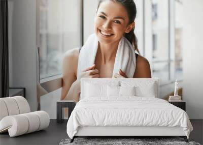 Relaxing. Portrait of happy cheerful woman in sportswear with white towel on her shoulders looking at camera with smile while standing in front of windows at gym Wall mural