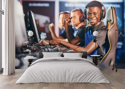 Side view of a cheerful african guy, professional cybersport gamer wearing headphones looking at camera and smiling while participating with team in eSport tournament Wall mural