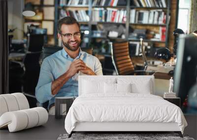 Smart and successful. Young and cheerful bearded man in eyeglasses and formal wear working on computer and smiling while sitting in modern office Wall mural