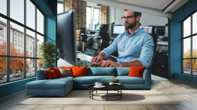 So busy. Young bearded man in eyeglasses and formal wear typing something on computer while sitting in the modern office Wall mural