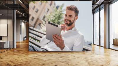 Social life. Young smiling man sitting on a bench in the park, holding his tablet. Rest, communication and relax concept Wall mural