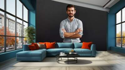 Successful project manager. Cheerful and young bearded man looking at camera with smile and keeping arms crossed while standing against dark background Wall mural