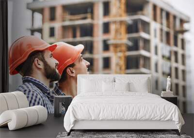 Teamwork. Two young and cheerful builders in red helmets are looking up and working at construction site Wall mural