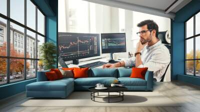 Trading online. Successful and young bearded trader in eyeglasses and formal wear working with laptop while sitting in his office in front of computer screens with trading charts Wall mural