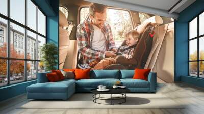 Traveling in the company of those we love is home in motion. Father fastening his baby boy that sitting in a car in safety chair. Family road trip Wall mural