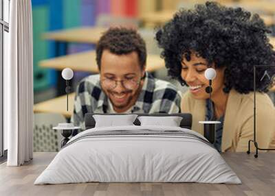 Two young happy diverse colleagues looking at laptop and discussing something while sitting at the desk in coworking space Wall mural