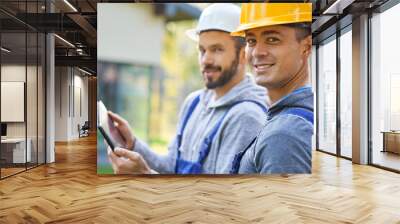 We will get it done. Portrait of two young male builders smiling at camera, using digital tablet pc while working at cottage construction site Wall mural