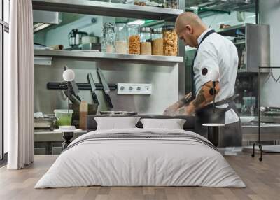 Working process. Back view of male chef in apron and with tattoos on his arms cutting a meat while standing in a restaurant kitchen. Wall mural