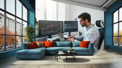 Workplace of trader. Young bearded trader wearing eyeglasses using his laptop while sitting in office in front of computer screens with trading charts and financial data Wall mural