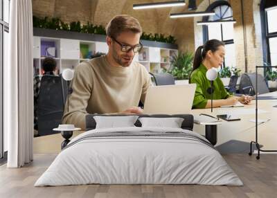 Young focused caucasian man wearing eyeglasses working on laptop while sitting at desk with his female colleague in the modern coworking space Wall mural