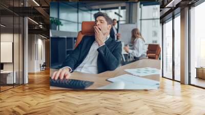 Young overtired businessman yawning while working inside modern office with computer Wall mural