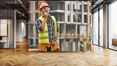 Сhecking work. Professional young builder in working uniform and red protective helmet talking to crane operator by walkie talkie while standing at construction site. Wall mural