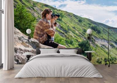 A young man with curly hair taking shots using camera in the nature, sitting on a rocky hill's slope Wall mural