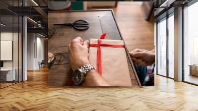 Young adult man with long blonde hair wrapping christmas present with red ribbon seated on wooden table in cozy apartment indoor. Wall mural