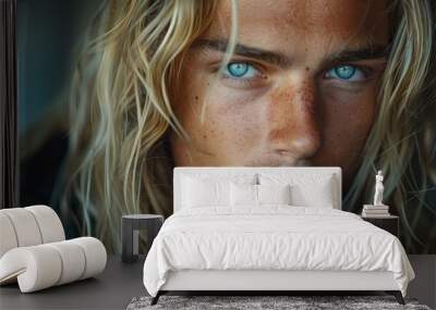 Close-up portrait of a young man with long blonde hair and striking blue eyes indoors at sunset Wall mural