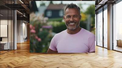 A smiling man in a pink shirt stands in front of a blurred background of flowers and a wooden fence. Wall mural