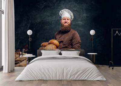 A bearded baker wearing a uniform holding fresh bread, standing next to a table, decorated with delicious bread loaves, baguettes and muffins in a dark studio Wall mural