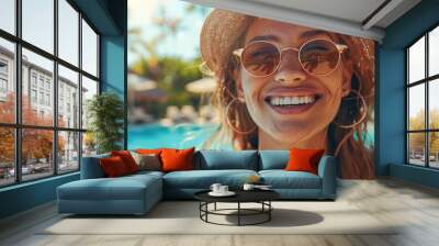 A beautiful young woman wears stylish sunglasses and a straw hat, with a resort pool in the background Wall mural