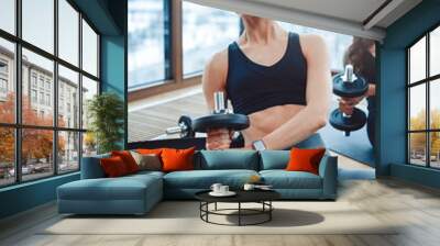 A muscular athletic young woman trains a press with dumbbells in her hands dressed in black top and leggins, sitting on a rug on the floor, in the background can see skyscraper reflection and city. Wall mural