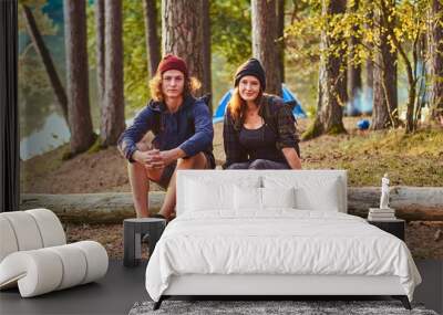 Attractive happy couple is relaxing in the green sunny forest while sitting on the log. There are campsite at background. Wall mural