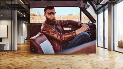 Bearded male in sunglasses dressed in brown leather jacket driving a retro car. Wall mural