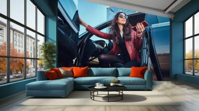 Beautiful smart women is posing in her new car while chatting on mobile phone. She is wearing red leather jacket and sunglasses. Wall mural