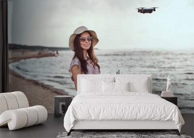 Charming girl in sunglasses and hat wearing dress enjoy the vacation on the beach, playing with the quadcopter. Wall mural