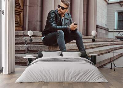 Fashionable guy dressed in a black jacket and jeans holds the smartphone sitting on steps against an old building in Europe. Wall mural