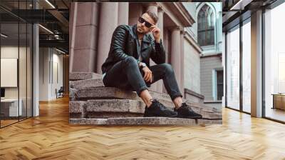 Fashionable guy dressed in a black jacket and jeans holds the smartphone sitting on steps against an old building in Europe. Wall mural