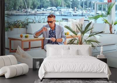 Fashionable successful man with stylish hair dressed in modern elegant clothes holds a cup of coffee while sitting on a sofa at outdoor cafe against the background of the city wharf. Wall mural