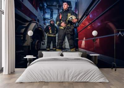 Full-length portrait of two brave firemen in protective uniform standing between two fire engines in the garage of the fire dept Wall mural