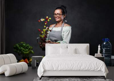 Happy middle-aged female chef tossing fresh fruit in the air from a blender in kitchen. Homemade healthy fruit smoothie. Studio photo on a dark background Wall mural